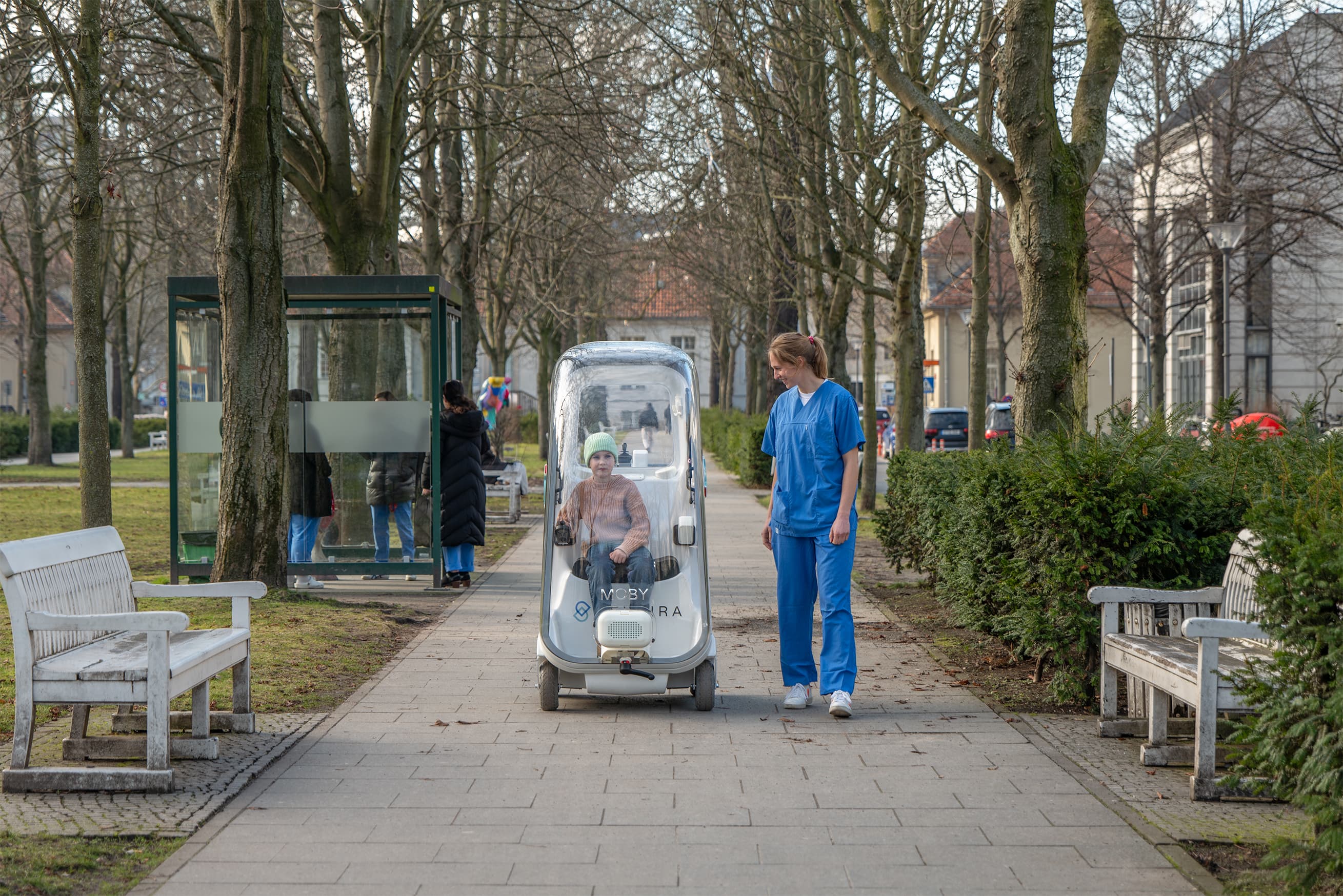 A child drives MOBY along a tree-lined hospital path, a nurse walking beside