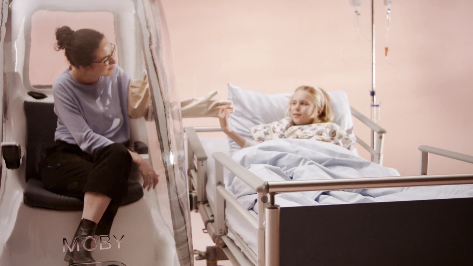 A relative sits in MOBY beside a girl's hospital bed and reaches through the glove port to her hand