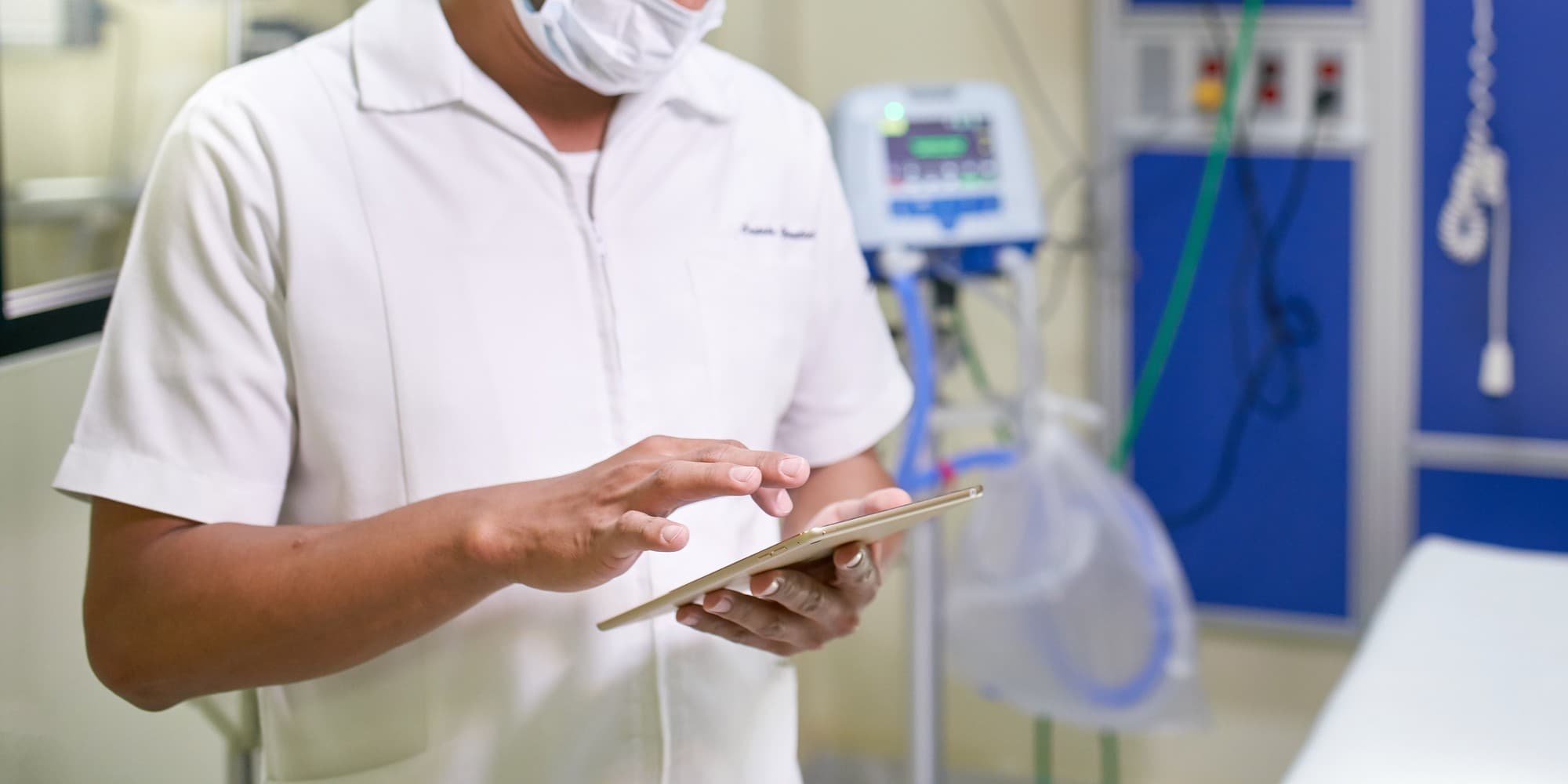 A clinician in a white coat and mask checks a tablet beside a ventilator in a hospital room
